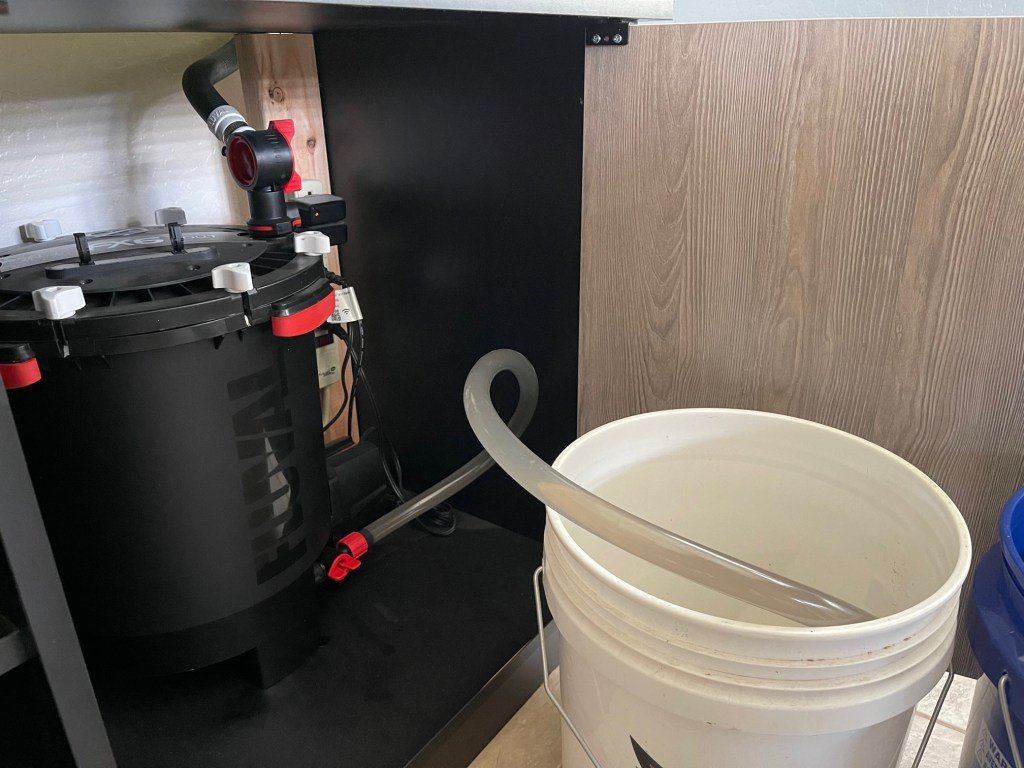 Filtro externo tipo canister instalado bajo el mueble de un acuario, junto a un cubo para el cambio de agua